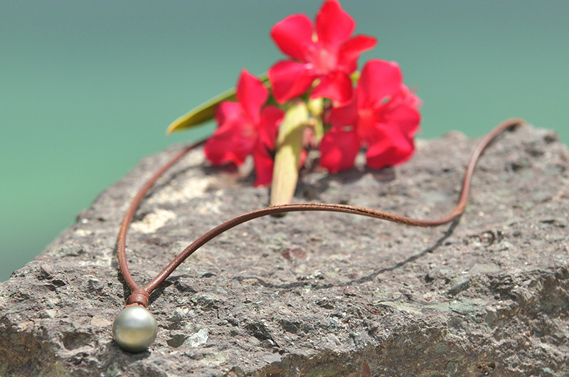 Unisex brown leather pendant necklace with two Tahitian cultured pearls displayed on a rock with red flowers in the background
