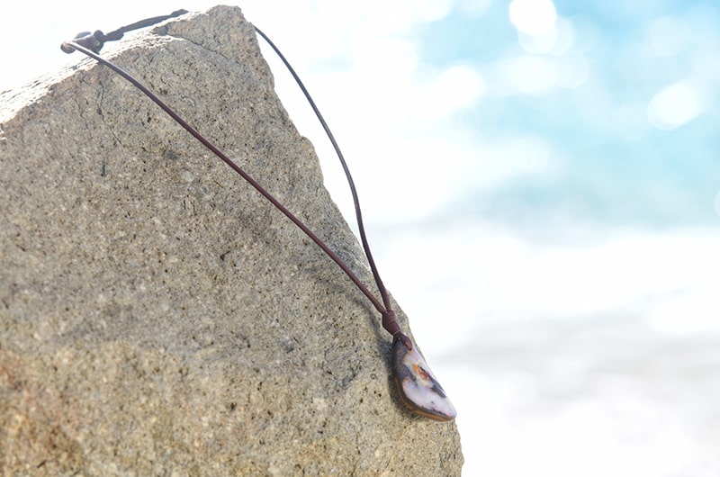 Full view of a brown leather necklace with an Australian opal pendant displayed on a rock by the sea