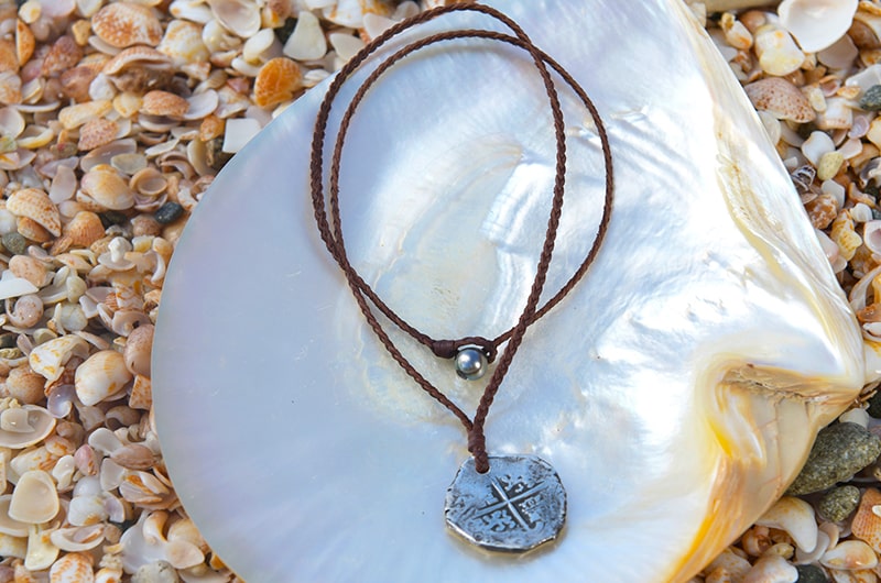 Braided leather pendant necklace with an antique silver eight reales coin and a Tahitian pearl displayed on mother-of-pearl