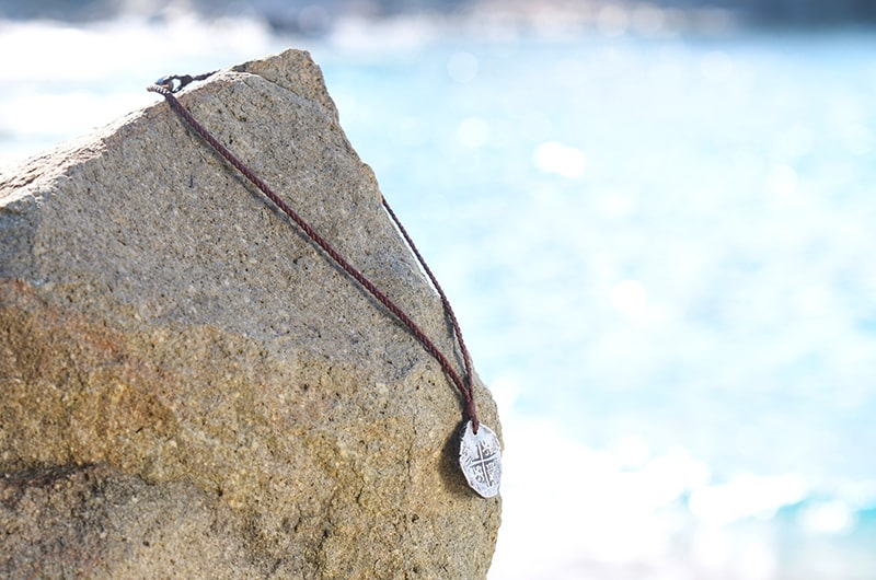 Braided leather pendant necklace with an antique silver eight reales coin displayed on a rock by the sea