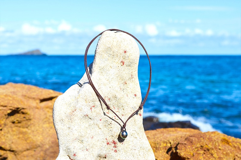 Vue d’ensemble du collier pendentif ajustable sur cordon marron avec perle de Tahiti de 10,5 mm présenté sur un rocher face à la mer