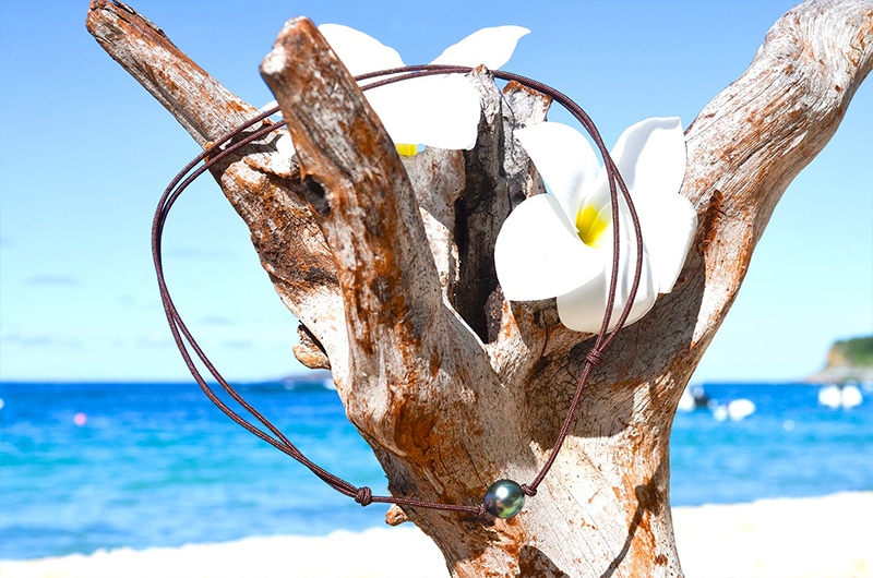 Full view of the adjustable choker necklace with a 9.5 mm semi-round Tahitian pearl displayed on driftwood by the sea