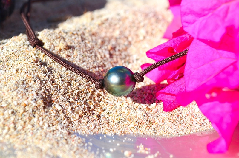 Detail of the 9.5 mm semi-round Tahitian cultured pearl and brown cord setting displayed on sand with pink flowers