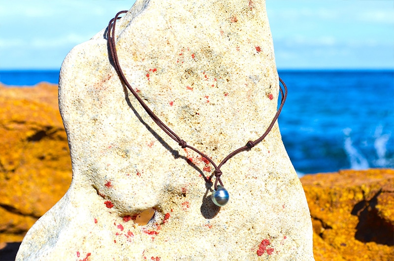 Vue d’ensemble du collier pendentif ajustable avec une perle de Tahiti cerclée de 10 mm présenté sur une roche face à la mer