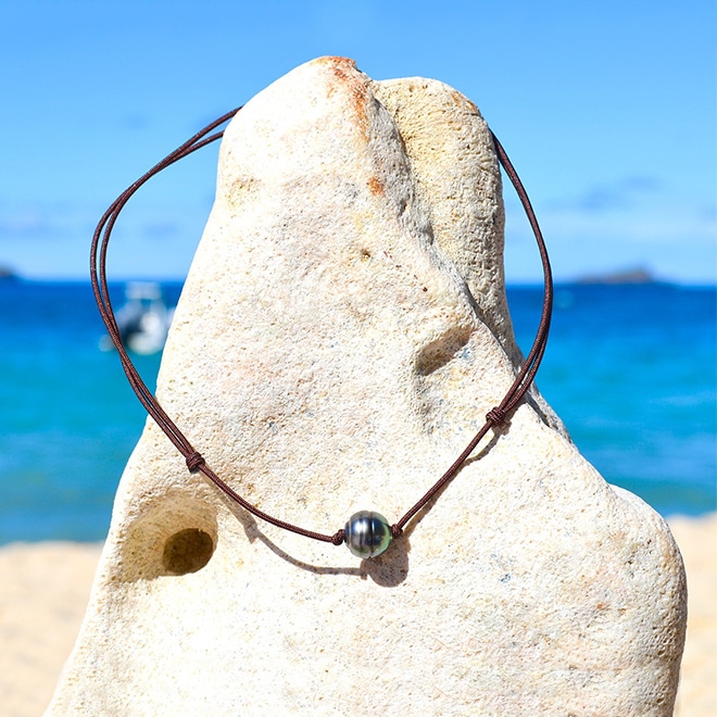 Collier ras du cou ajustable sur cordon marron avec une perle de culture de Tahiti de 11 mm présenté sur une roche face à la mer