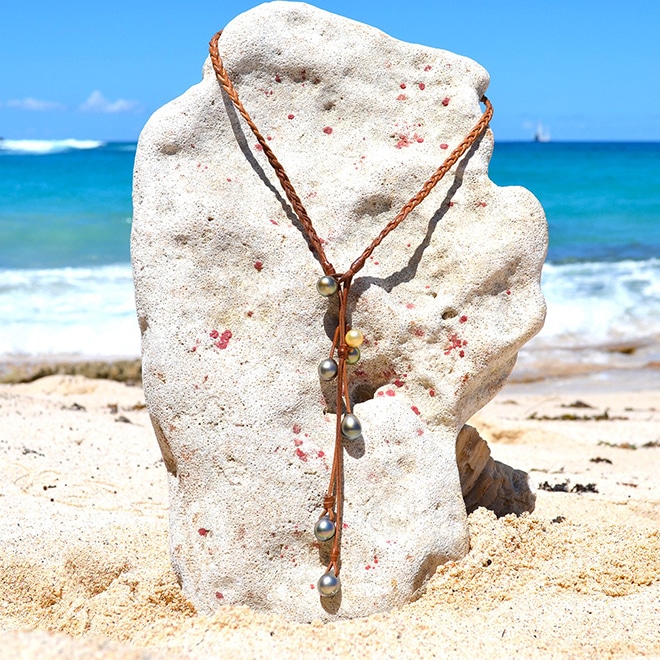 Collier en cuir tressé avec six perles de culture de Tahiti et une perle dorée des mers du Sud présenté sur un support corallien à la plage