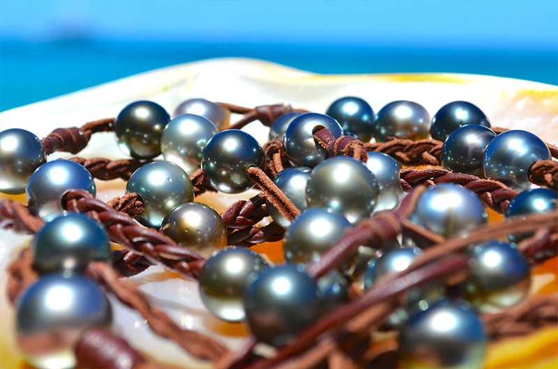 Gros plan sur les perles de culture de Tahiti et le cuir tressé du sautoir disposés sur une nacre face à la mer