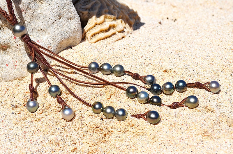 Sautoir en cuir tressé avec 33 perles de culture de Tahiti déployé sur le sable près d’un support corallien