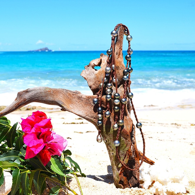 Vue d’ensemble du sautoir en cuir tressé avec 33 perles de culture de Tahiti présenté sur du bois flotté à la plage