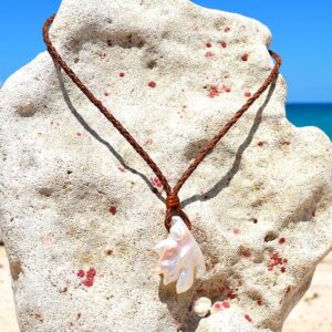 Collier pendentif en cuir tressé avec une grande perle de culture d’eau douce baroque présenté sur un support corallien face à la mer