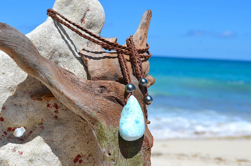 Vue d’ensemble du collier pendentif tressé avec un Larimar et quatre perles de Tahiti présenté sur du bois flotté à la plage