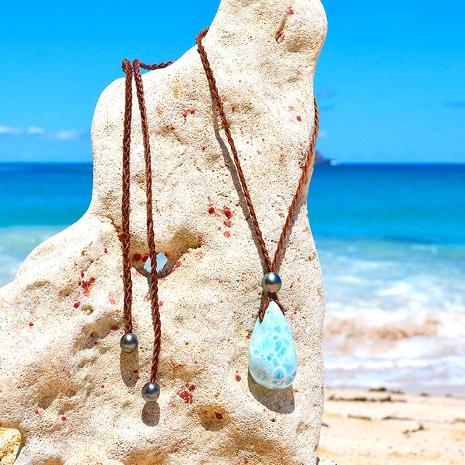 Collier pendentif en cuir tressé avec un Larimar et quatre perles de culture de Tahiti présenté sur un support corallien face à la mer