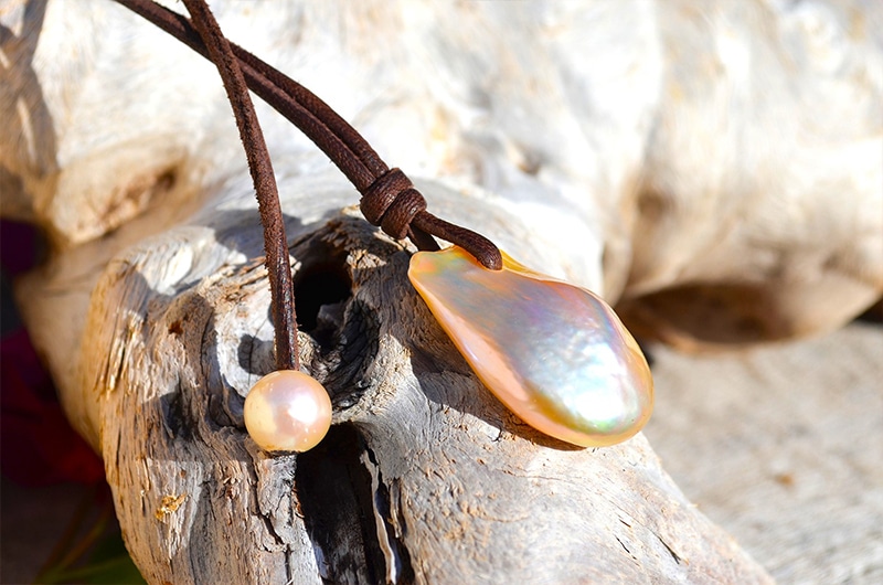 Alternate view of the large Fireball pearl, pink freshwater pearl and brown leather on the necklace displayed on driftwood