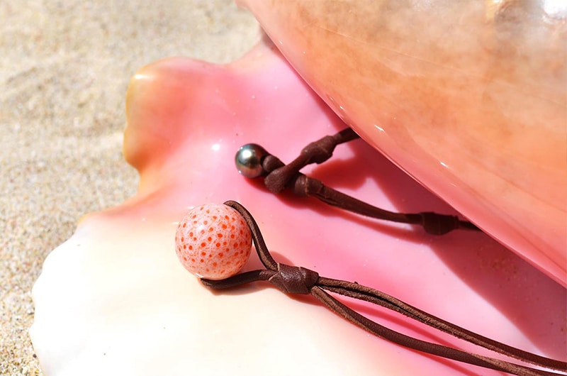Close view of the coral element, Tahitian pearl and brown leather on the pendant necklace displayed on a pink seashell