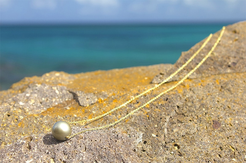 Collier en chaîne d’or 14 carats avec une perle de culture de Tahiti de 13 mm déployé sur une roche face à la mer