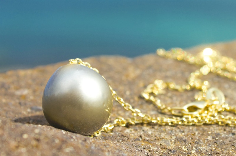 Gros plan sur la perle ronde de culture de Tahiti de 13 mm et la chaîne en or 14 carats du collier posées sur une roche