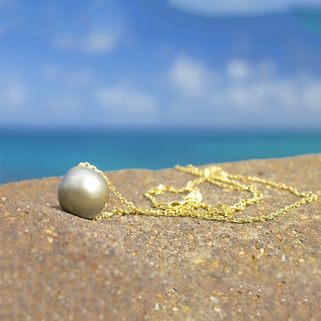 Collier en chaîne d’or 14 carats avec une perle ronde de culture de Tahiti de 13 mm présenté sur une roche face à la mer