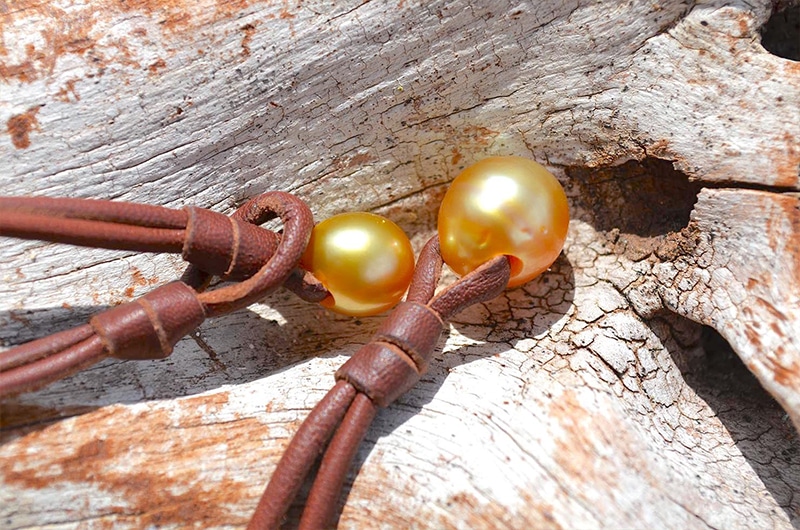 Détail des deux perles de culture dorées des mers du Sud et du montage en cuir du collier sur du bois flotté