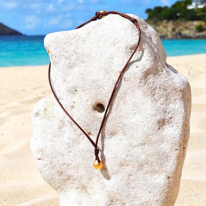 Collier pendentif en cuir avec deux perles de culture dorées des mers du Sud présenté sur un support corallien à la plage