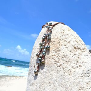 Vue d’ensemble du collier en cuir tressé avec 25 perles de culture de Tahiti présenté sur un support corallien à la plage