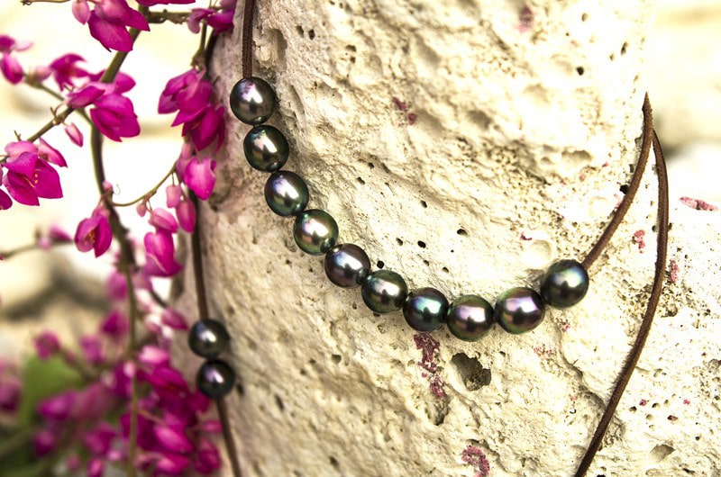 Long necklace with 50 semi-baroque Tahitian cultured pearls displayed vertically against a rock among pink flowers
