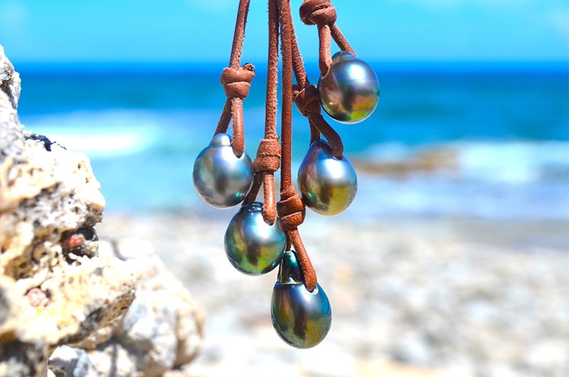 Vue rapprochée de la grappe de cinq perles de culture de Tahiti et des nœuds en cuir marron du collier face à la mer