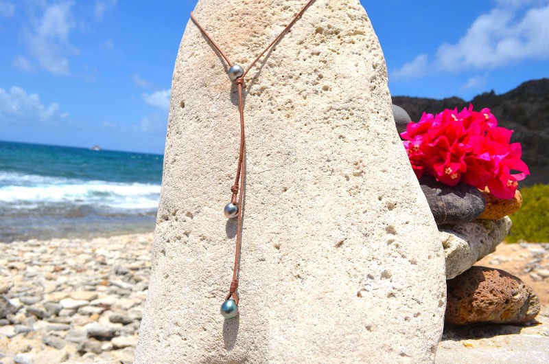 Vue d’ensemble du sautoir ajustable en cuir avec trois perles de Tahiti semi-baroques présenté sur un support corallien à la plage