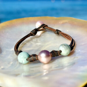 Bracelet en cuir marron avec billes d’émeraude et perle de culture blanche présenté sur une nacre face à la mer