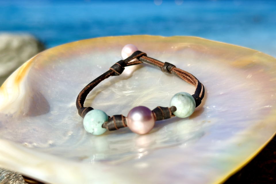 Brown leather bracelet with emerald beads and a white cultured pearl displayed on mother-of-pearl facing the sea