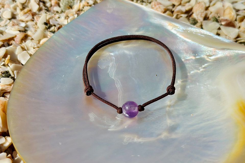 Adjustable brown cord bracelet with an 8 mm amethyst displayed on mother-of-pearl