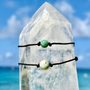 Deux bracelets ajustables sur cordon noir avec émeraudes de différentes tailles présentés sur un cristal face à la mer