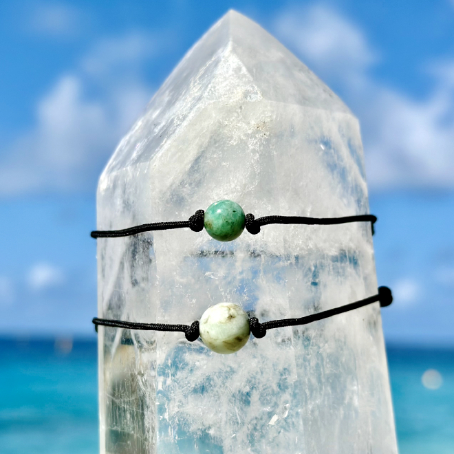 Deux bracelets ajustables sur cordon noir avec émeraudes de différentes tailles présentés sur un cristal face à la mer