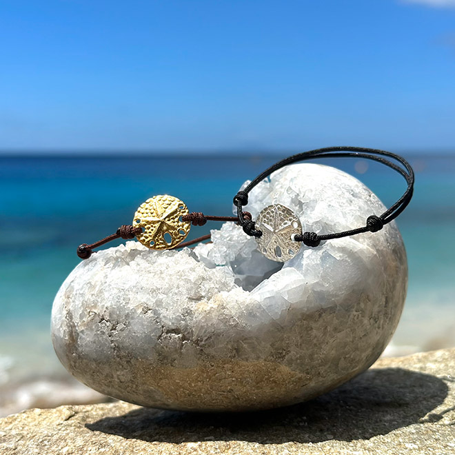 Adjustable bracelets with a sterling silver or gold-finished sand dollar medal displayed on a rock facing the sea