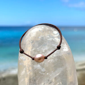 Bracelet ajustable sur cordon marron avec une perle d’eau douce ovale rose présenté sur un cristal face à la mer