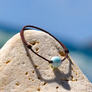 Bracelet ajustable sur cordon marron avec une bille de Larimar de 10 mm présenté sur une roche claire face à la mer