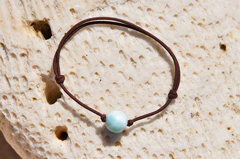 Adjustable brown cord bracelet with one Larimar bead displayed on a pale stone