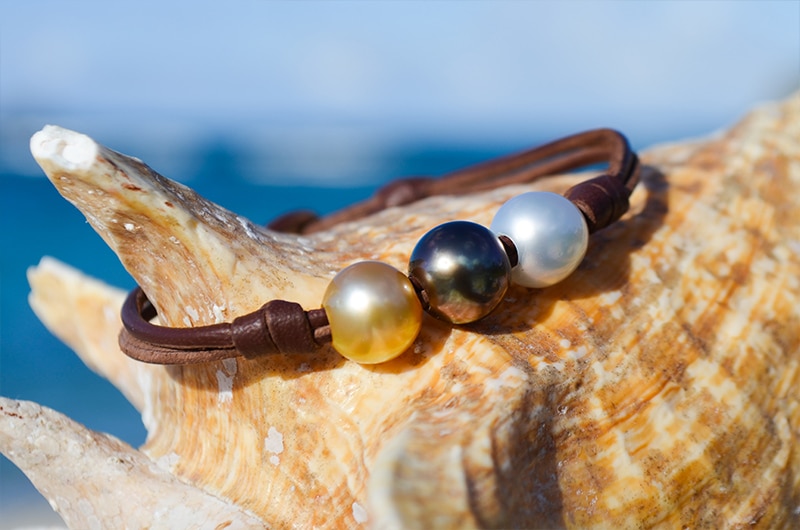 Leather bracelet with three multicolored pearls displayed on a seashell facing the sea