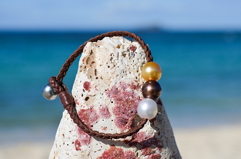Bracelet en cuir tressé avec trois perles multicolores présenté autour d’une roche claire face à la mer
