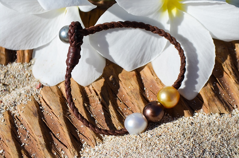 Bracelet tressé avec trois perles multicolores présenté sur le sable près de fleurs blanches