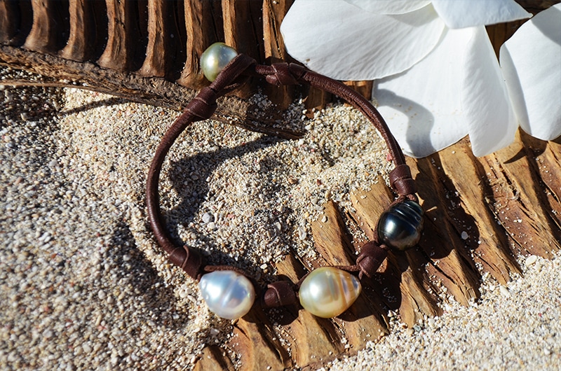 Full view of the bracelet with three multicolored circled pearls displayed on the sand near white flowers