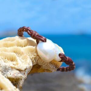 Bracelet en cuir marron tressé avec une perle de culture d’eau douce blanche présenté sur du corail face à la mer