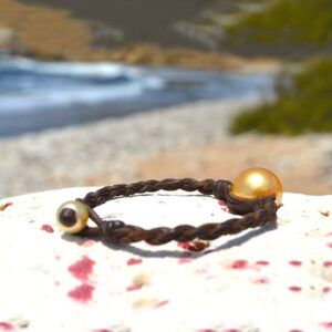 Bracelet en cuir marron tressé avec une perle de culture dorée d’Australie présenté sur une roche face à la plage