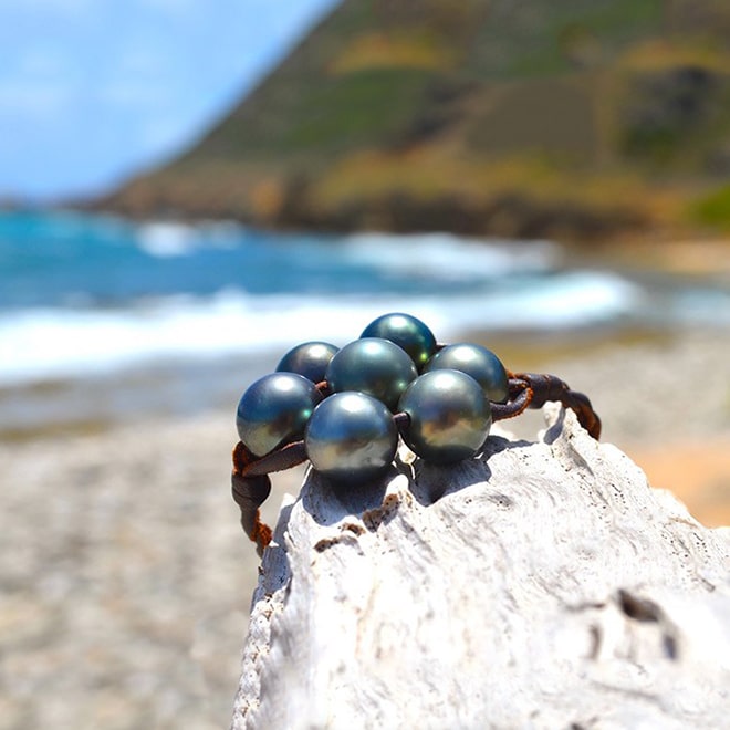 Bracelet en cuir marron tressé avec sept perles de culture de Tahiti présenté sur du bois face à la plage