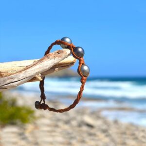 Bracelet en cuir marron tressé avec trois perles de culture de Tahiti présenté sur du bois face à la mer