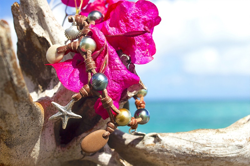 Bracelet avec perles de Tahiti, galets de rivière et breloques présenté près de fleurs roses face à la mer