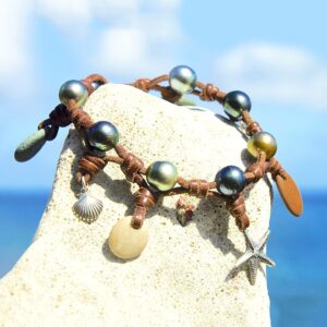 Bracelet en cuir avec perles de culture de Tahiti, galets de rivière et breloques présenté sur une roche face à la mer