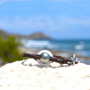 Bracelet en cuir marron tressé avec anneau de nacre, perle de culture de Tahiti et Keshi présenté sur une roche face à la mer