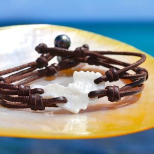 Bracelet en cuir marron avec un coquillage en forme de Saint-Barthélemy et deux perles de culture de Tahiti présenté sur une nacre face à la mer