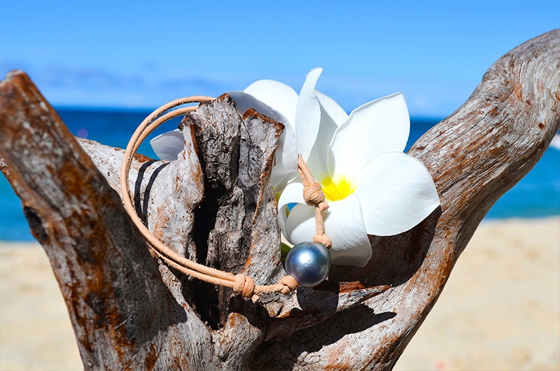 Bracelet ajustable en cuir marron avec une perle de culture de Tahiti cerclée de 13 mm présenté sur du bois avec une fleur blanche