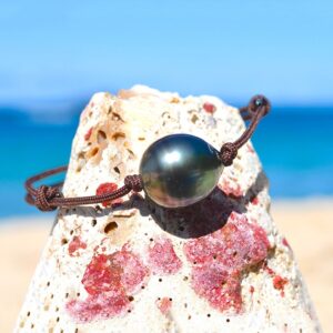 Adjustable brown cord bracelet with a 13 mm circled Tahitian cultured pearl displayed on a rock by the sea